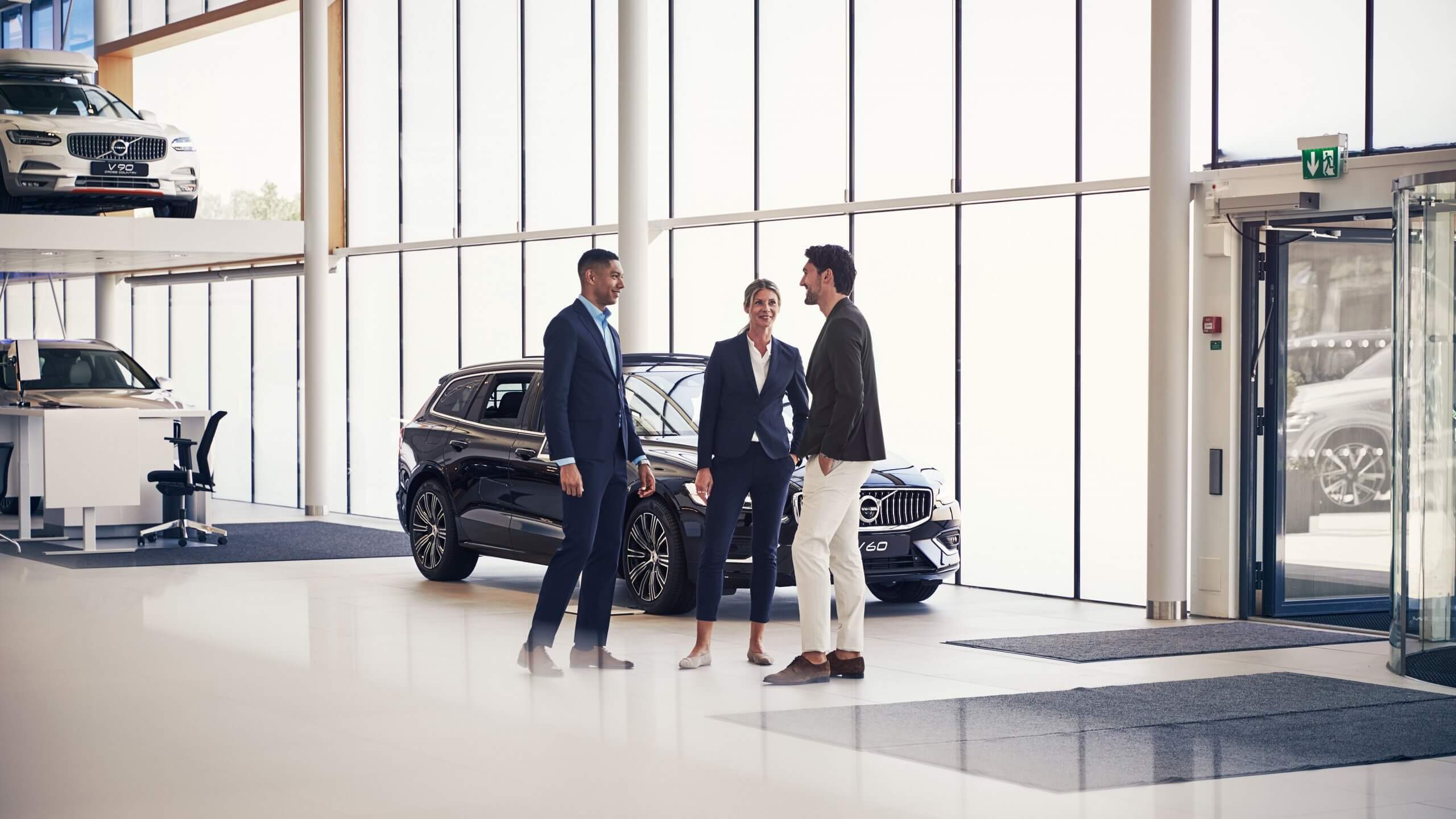 A Volvo dealer/retailer showroom in the background and a sales agent with custormers chatting.